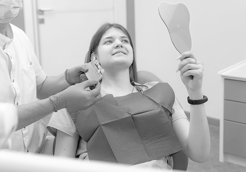 A patient viewing her smile in a mirror while a dentist holds veneer samples.
